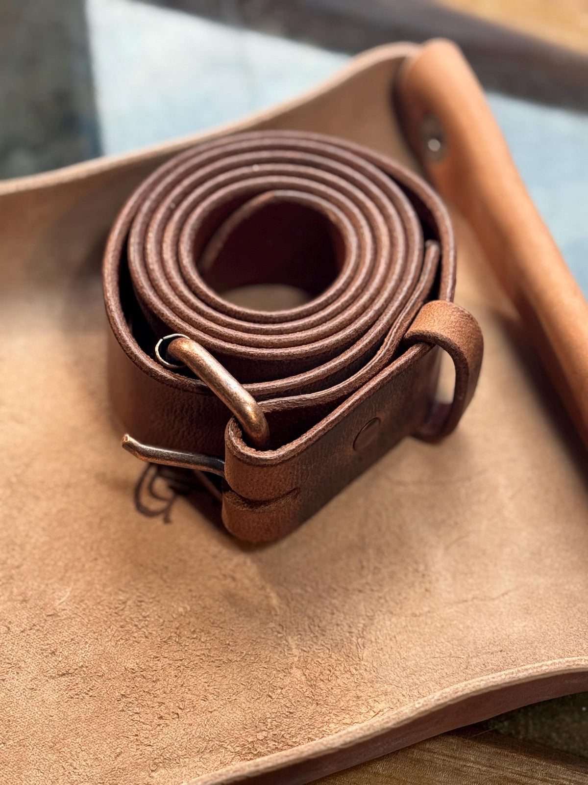 A coiled brown leather belt with a brass buckle resting on a tan leather pad, with a wooden tool partially visible in the background.
