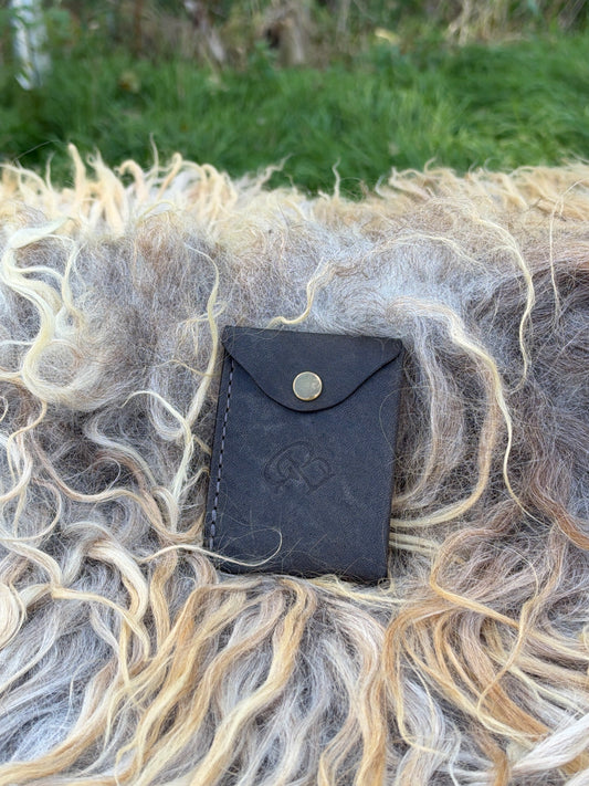 A small black leather card holder or wallet with a snap button closure, placed on a bed of curly wool in an outdoor setting with green grass in the background.