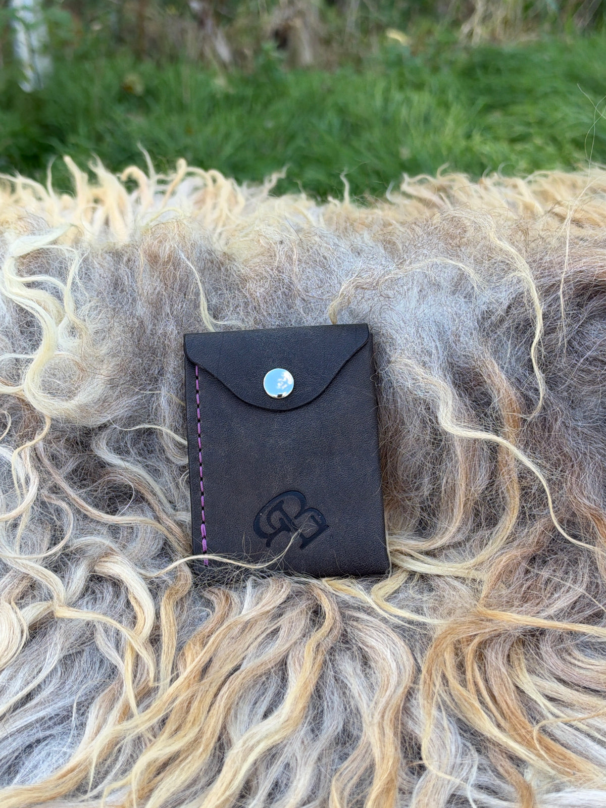 A small black leather card holder or wallet with a snap button closure, placed on a shaggy sheepskin or wool surface outdoors with blurred green grass in the background.