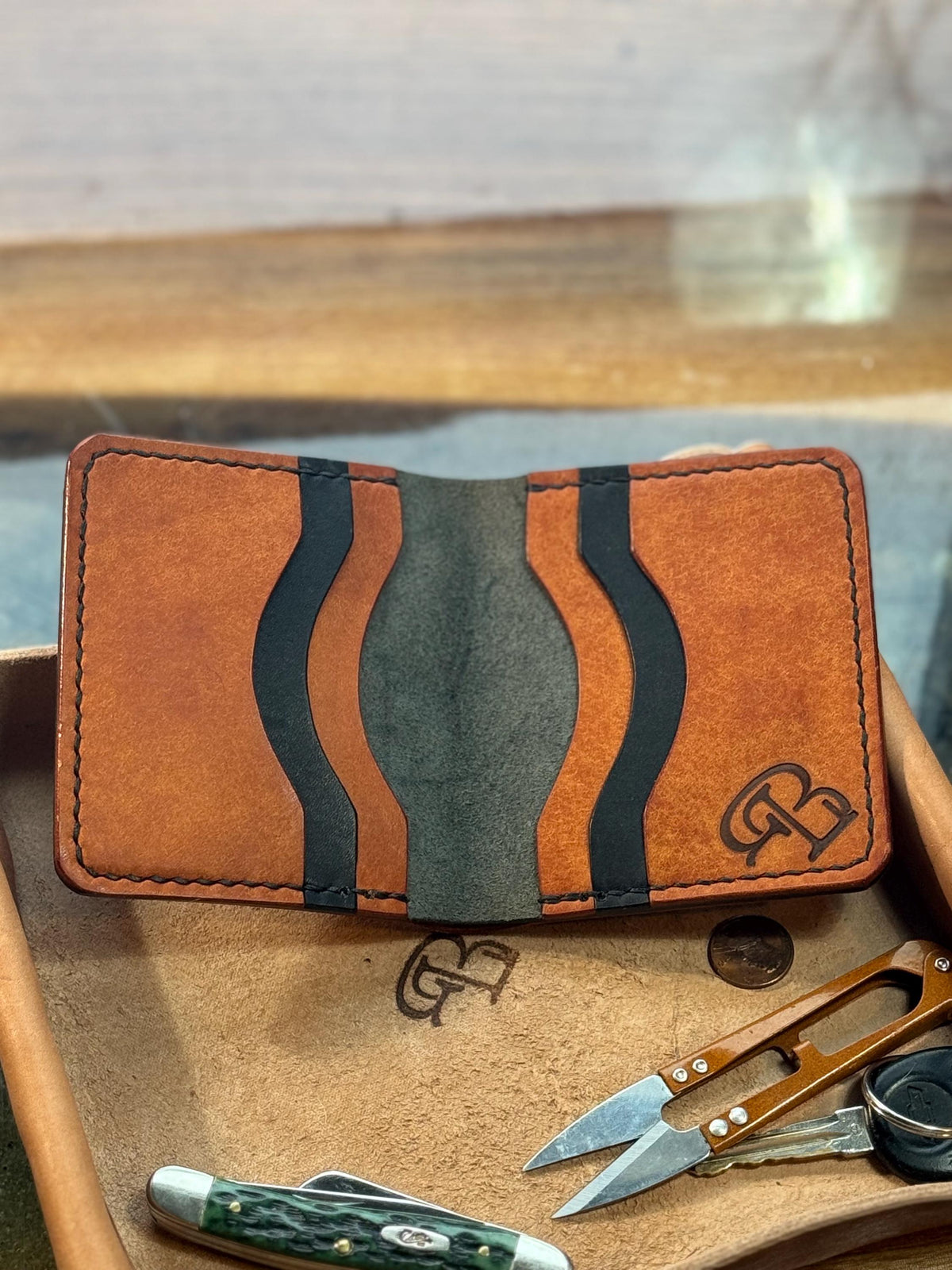 A brown leather card holder wallet with a wavy stripe pattern in black and dark gray, displayed on a wooden tray alongside a pocket knife, scissors, and keys.