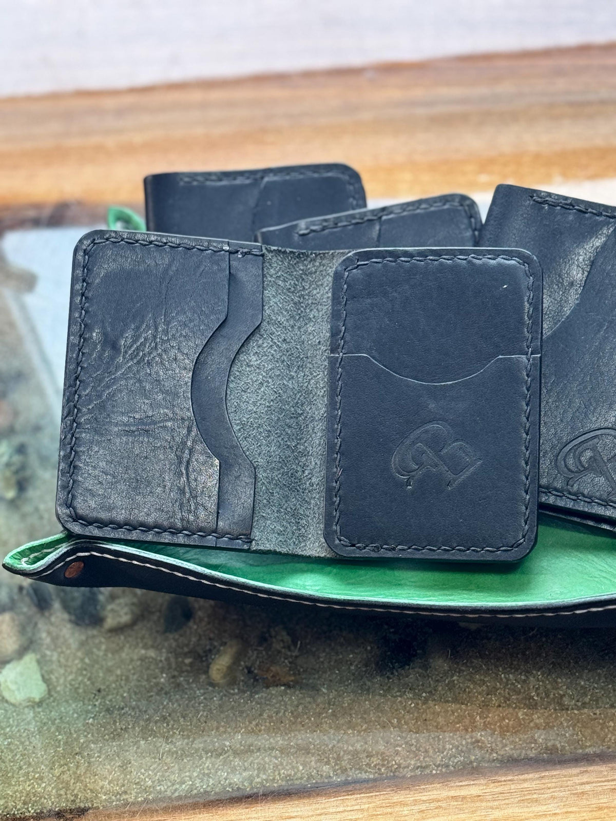 A close-up of a black leather card holder wallet with multiple card slots and a front slip pocket, resting on a green leather surface with other wallets in the background.