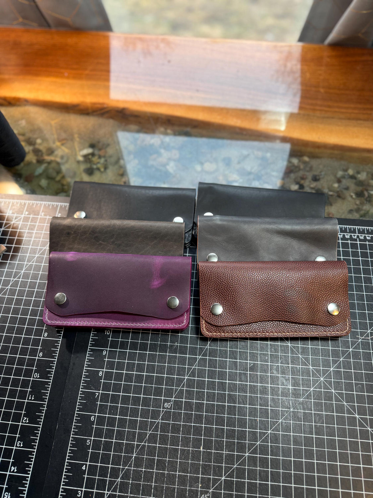 Four small leather card holders or wallet-style pouches arranged in two pairs on a black gridded cutting mat. The front row includes a purple pouch, a brown pebbled leather pouch, and two darker brown leather pouches behind them.