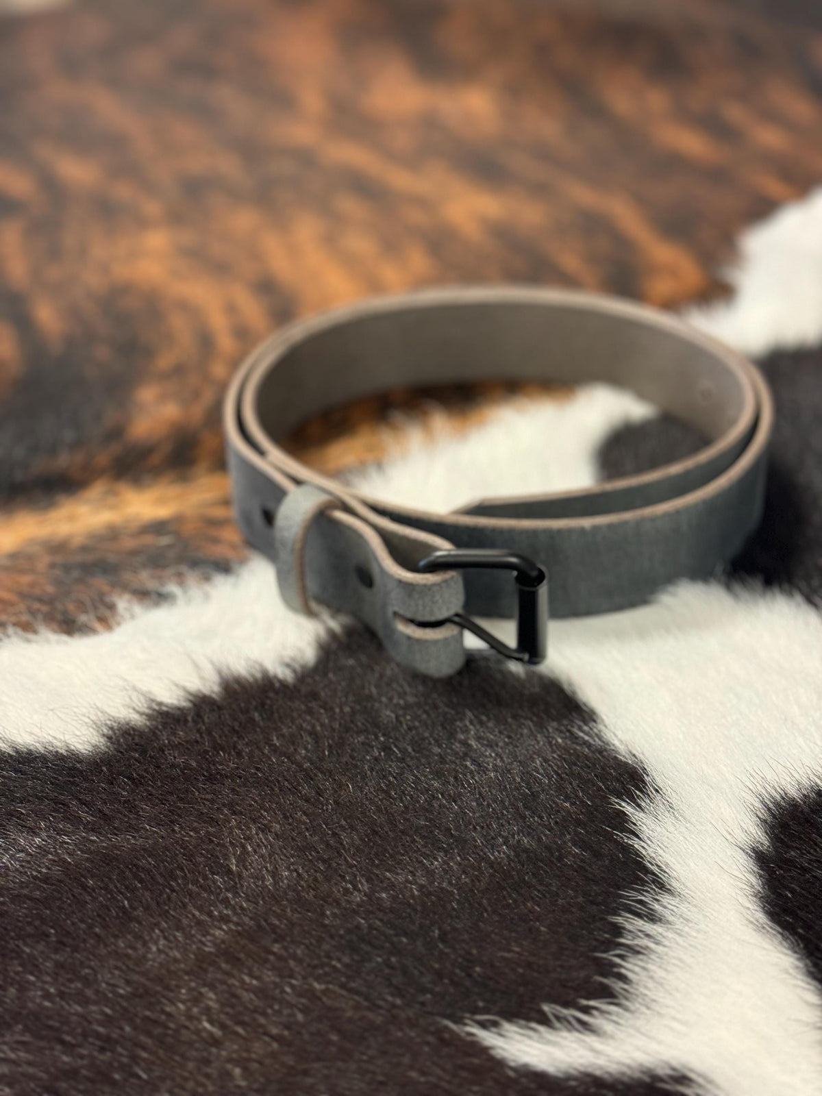 A gray leather belt with a black metal buckle, coiled on a black and white cowhide rug.