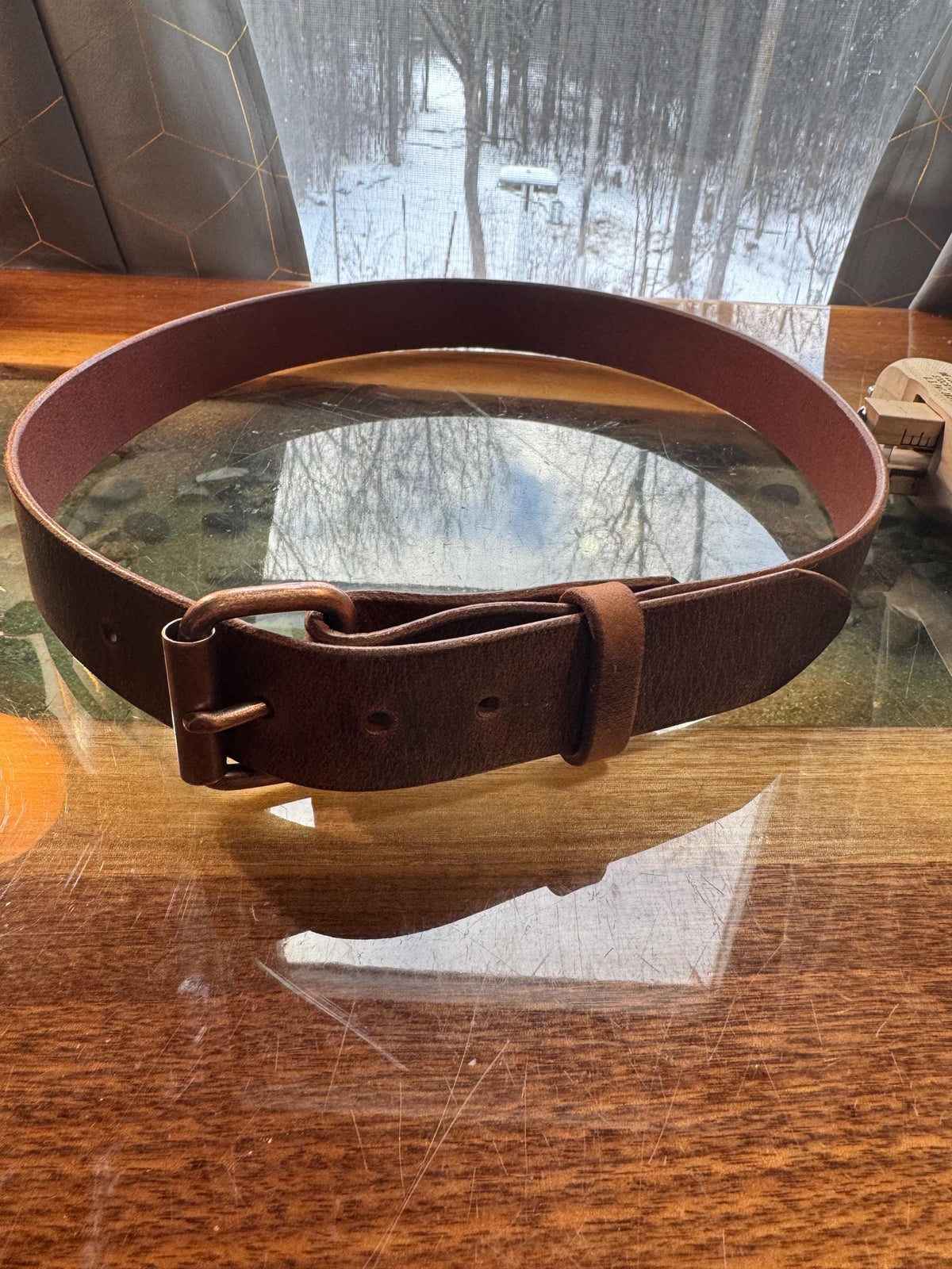 A dark brown leather belt with a matching metal buckle, resting on a reflective wooden table with a snowy outdoor scene visible in the background.