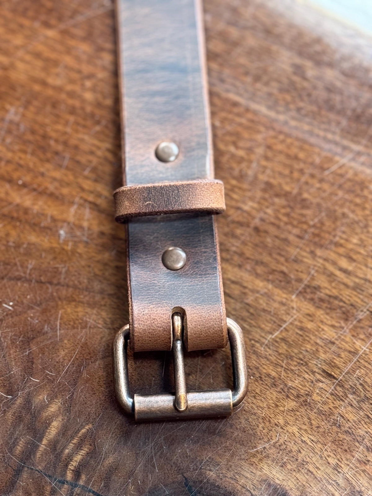 A close-up of a dark brown leather belt strap with an antique bronze buckle, laid diagonally on a textured wooden surface.