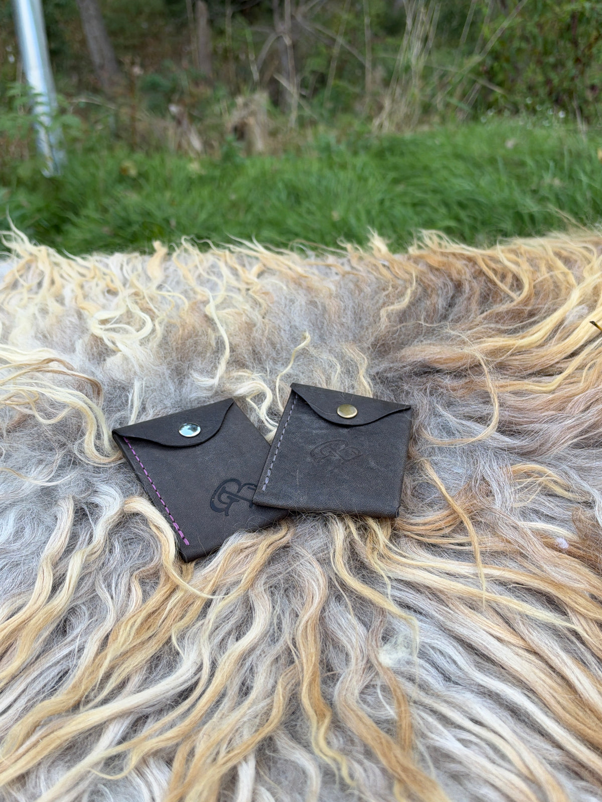 Two small black leather card holders or wallet-style pouches with snap closures, placed on top of a shaggy sheepskin or wool surface outdoors.