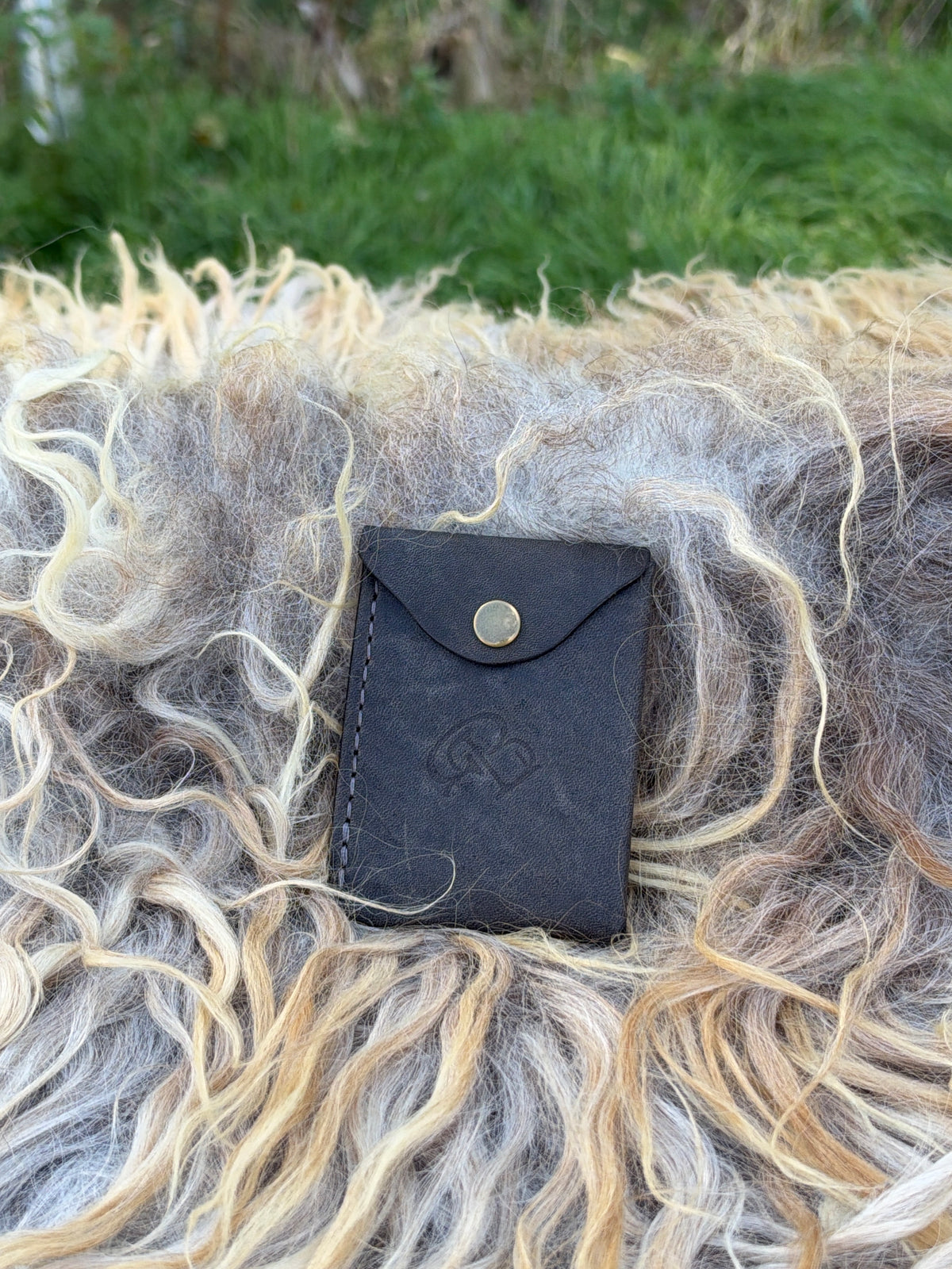 A small black leather card holder or wallet with a snap button closure, placed on a bed of curly wool in an outdoor setting with green grass in the background.