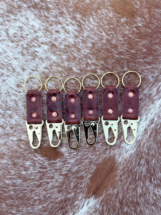 Six leather keychain clips arranged in a row on a brown and white cowhide background. The keychains feature reddish-brown leather bodies with brass key rings and clips, with three showing gold-toned metal and three showing silver-toned metal.