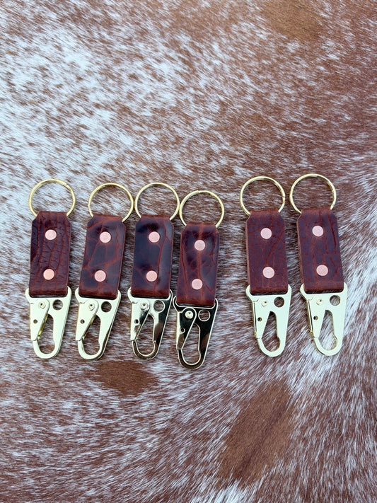 Five leather keychain clips arranged in a row on a brown and white cowhide background. Each clip features a reddish-brown leather tab with a pink stitched dot, a dark wood-grain pattern, and a gold-tone metal clasp and key ring.