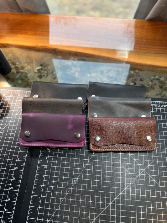 Four small leather card holders or wallet-style pouches arranged in two pairs on a black gridded cutting mat. The front row includes a purple pouch, a brown pebbled leather pouch, and two darker brown leather pouches behind them.