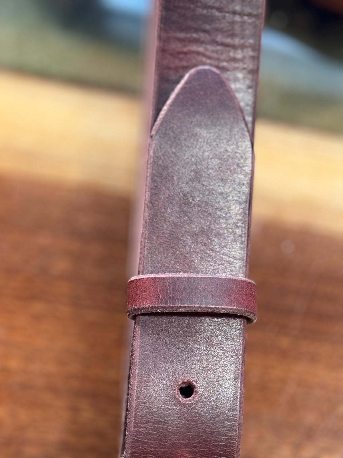 A close-up of a dark reddish-brown leather strap, likely a watch band or belt, showing the tip of the loop and the adjustment hole against a blurred wooden background.