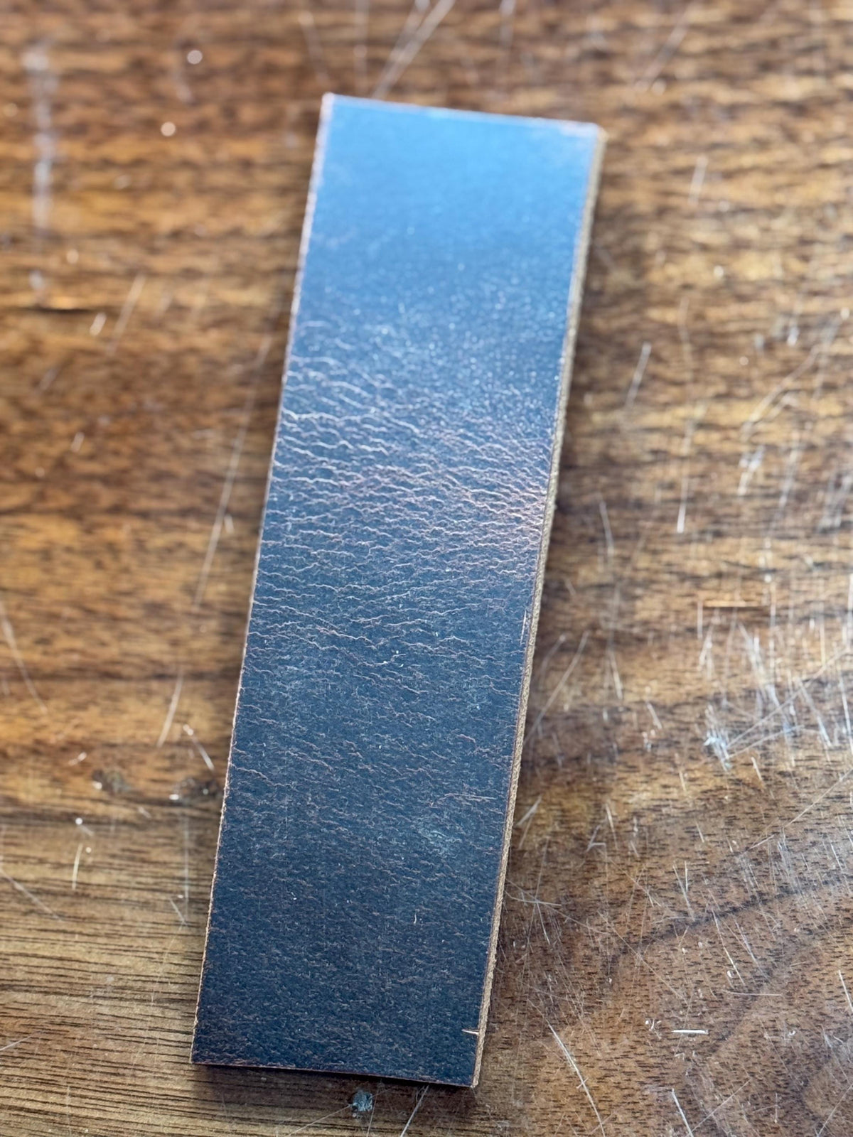 A rectangular black leather strap or bar with a textured surface, placed diagonally on a scratched wooden surface.
