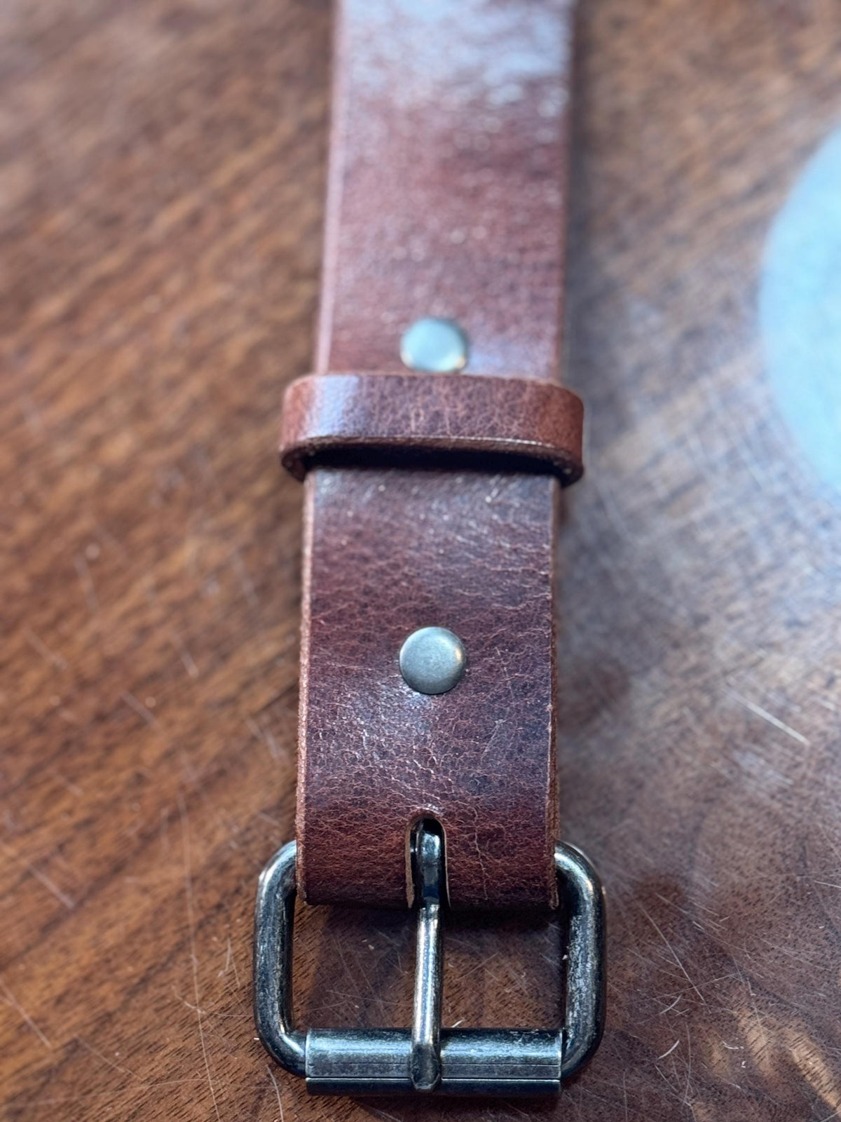 A close-up of a brown leather watch strap with a dark metal buckle, laid on a wooden surface.