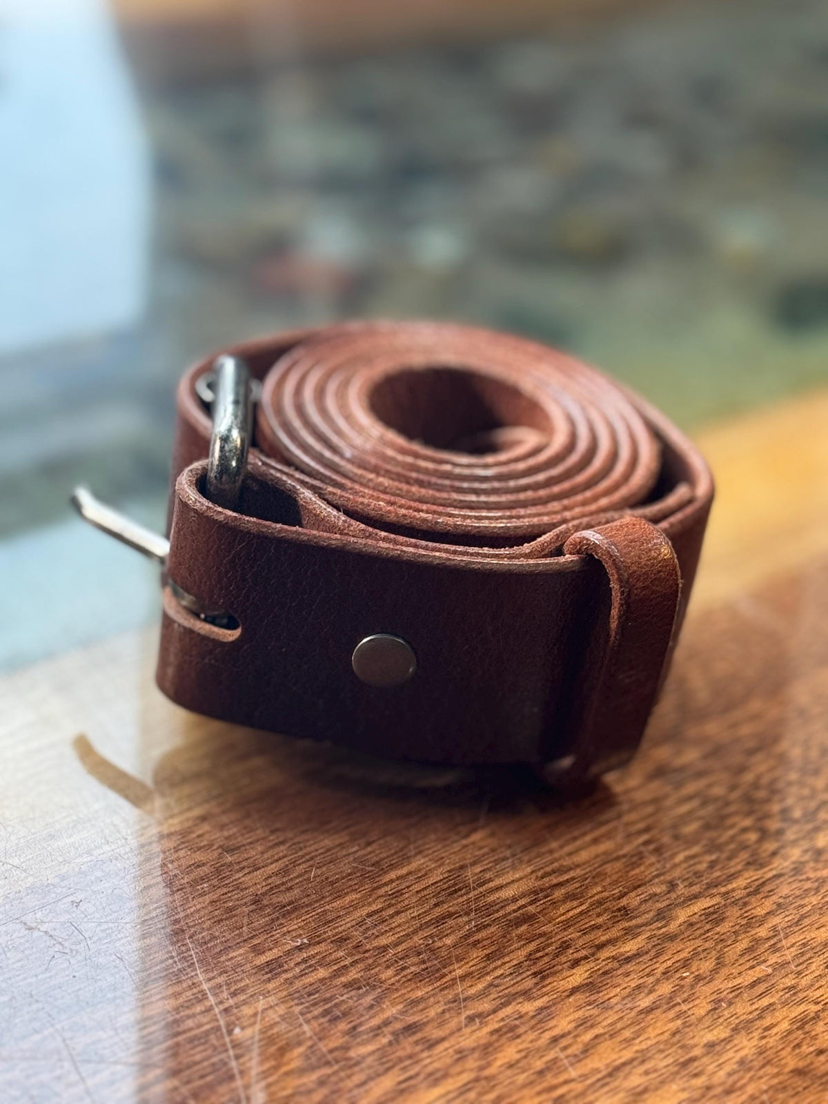 A rolled brown leather belt with a silver-tone metal buckle and rivet, resting on a polished wooden surface.