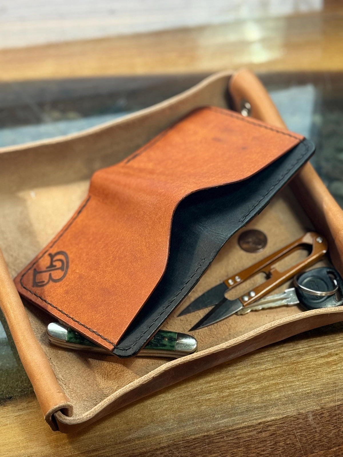 A tan leather card holder wallet with a black interior, shown open and resting on a leather tray with scissors and keys on a wooden surface.