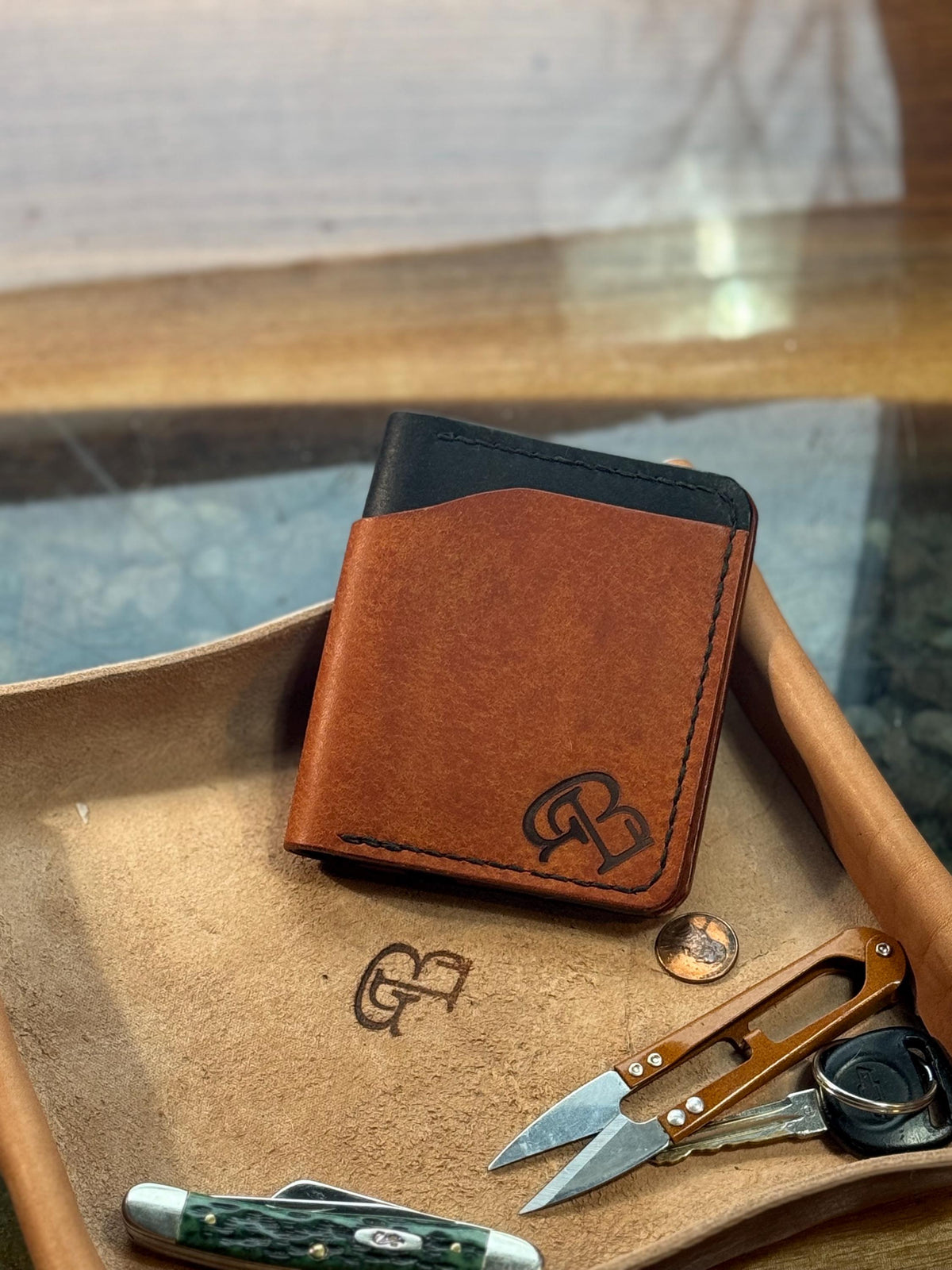 A brown leather card holder wallet with a black top panel and an embossed monogram logo, displayed on a tan leather tray with a pocket knife, scissors, and keys.