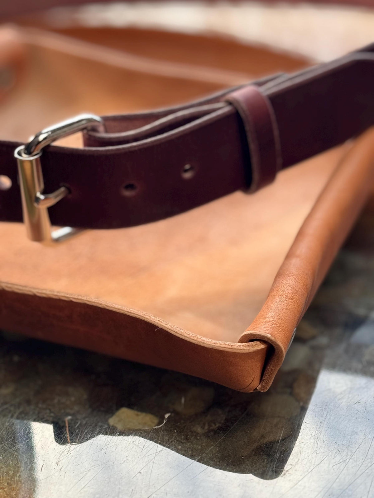 A close-up of a dark brown leather belt with a silver buckle resting on a tan leather tray or pad, set on a reflective dark surface.