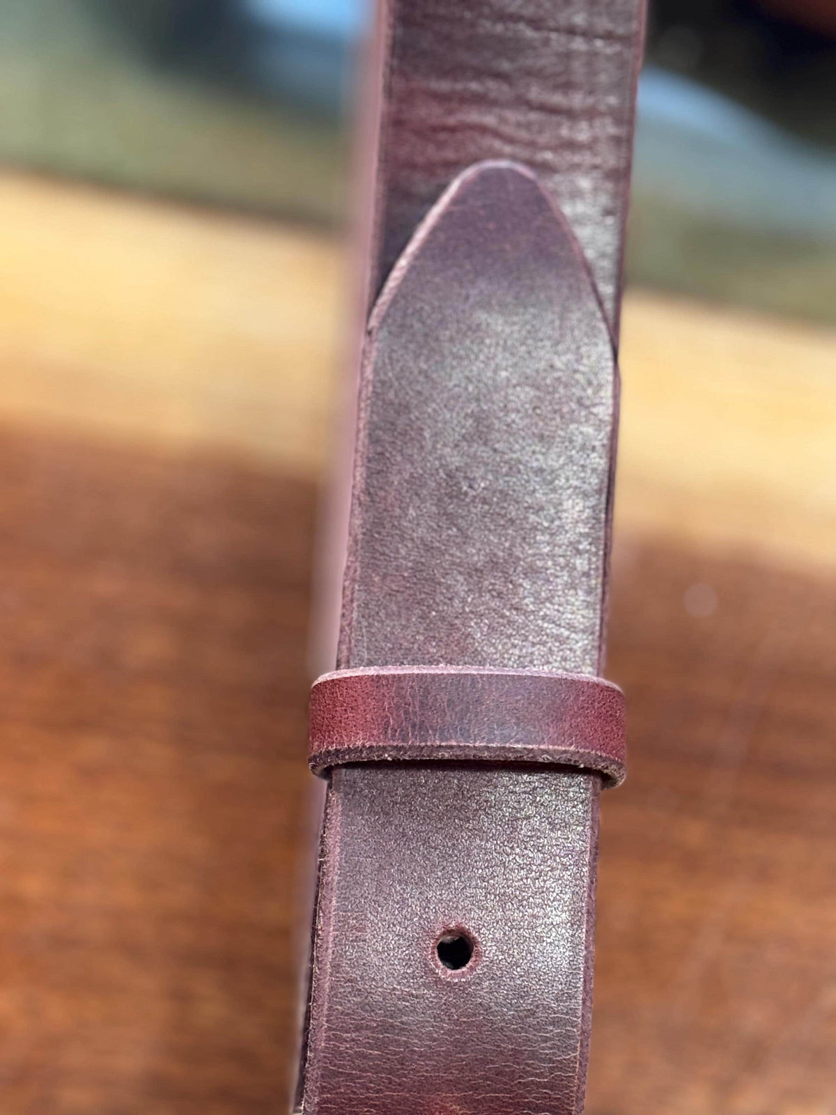 A close-up of a dark reddish-brown leather strap, likely a watch band or belt, showing the tip of the loop and the adjustment hole against a blurred wooden background.