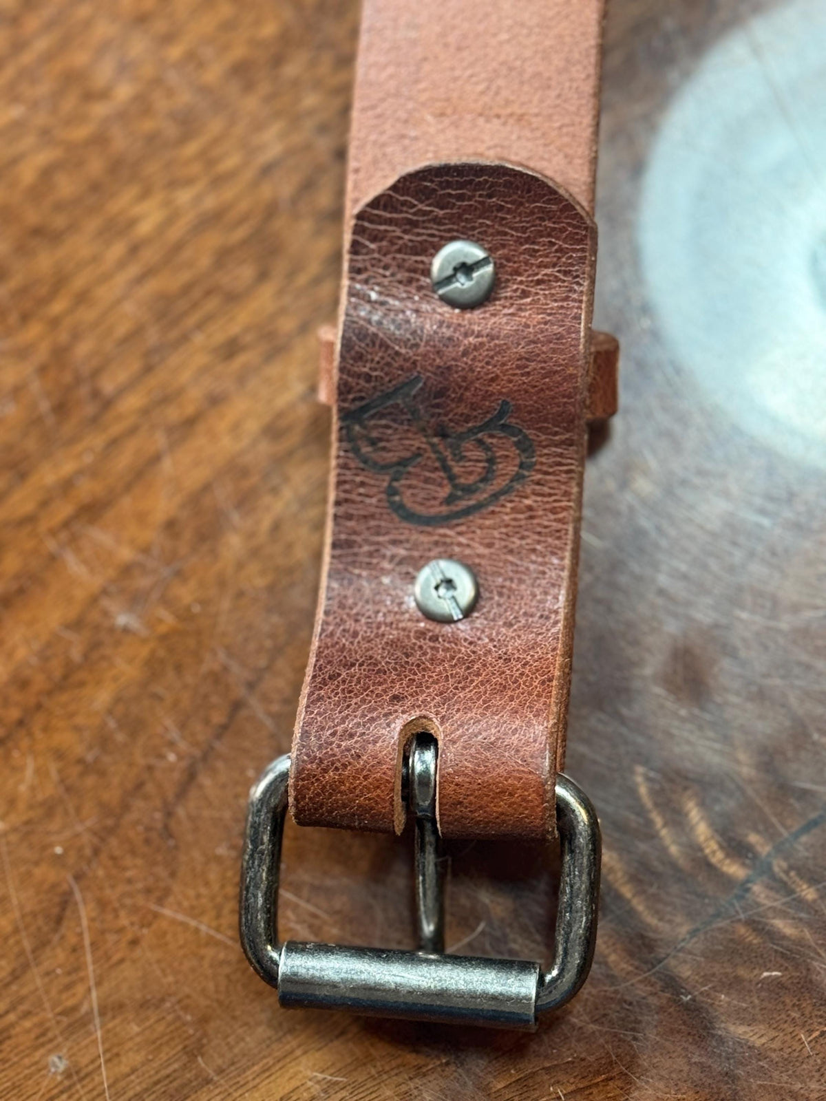 A close-up of a brown leather strap with a metal buckle, resting on a wooden surface. The leather shows a stamped logo and two silver screws.