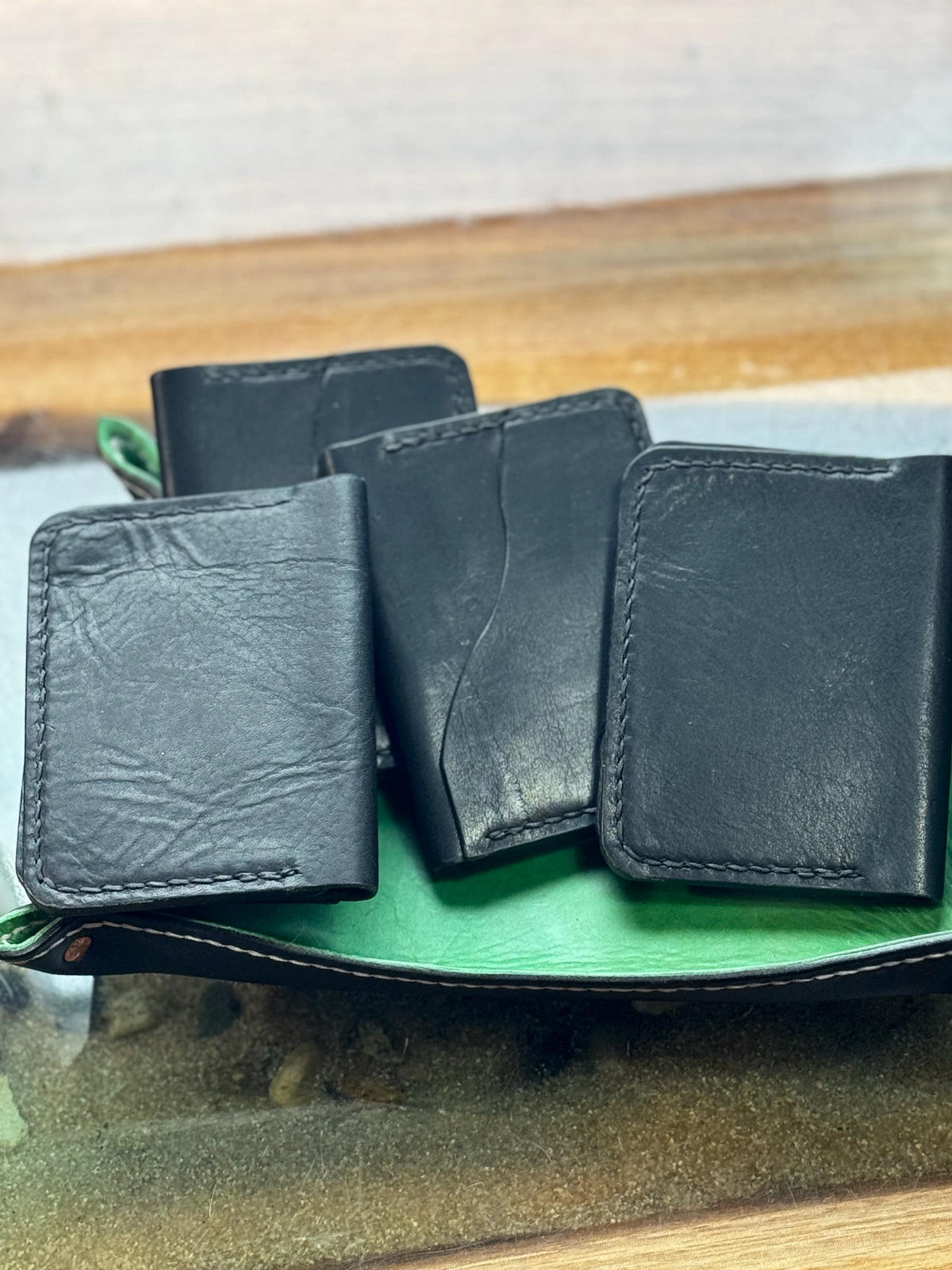Four black leather card holders or wallet-style accessories arranged on a green tray, photographed on a wooden surface with a light wall in the background.