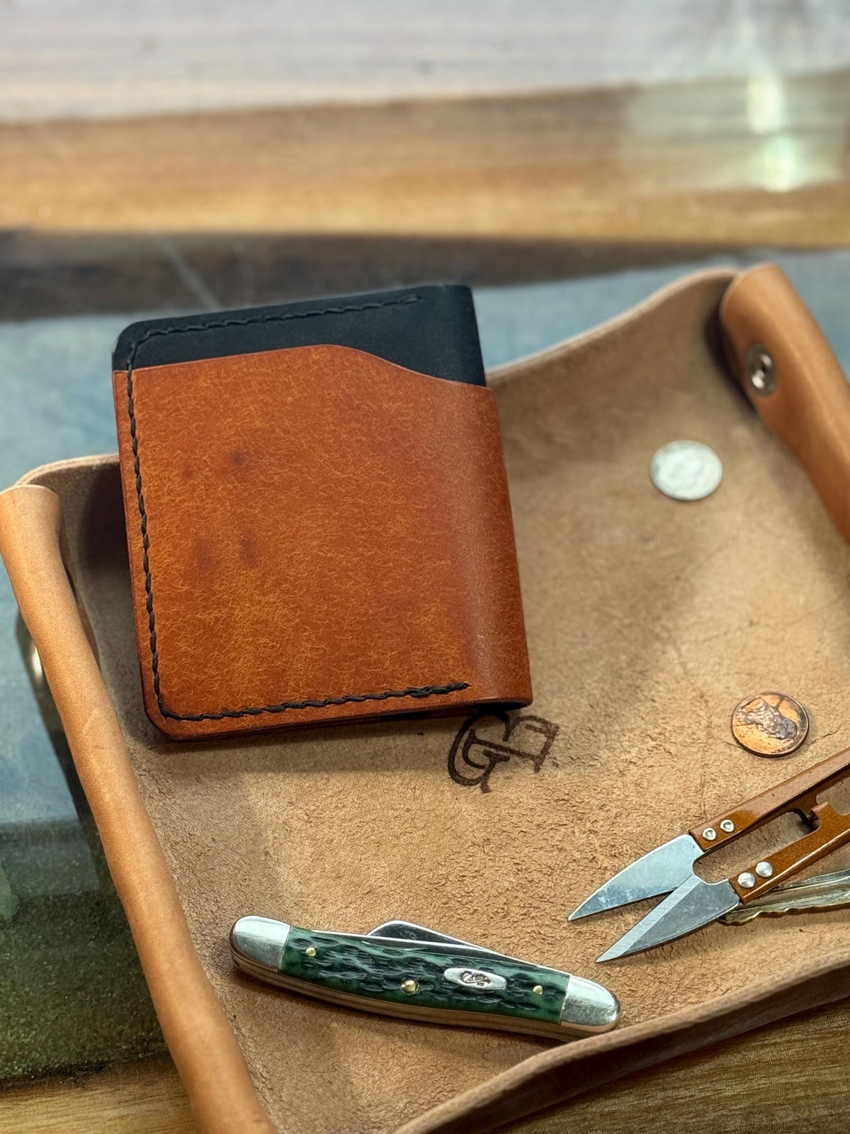A brown leather bifold wallet with a black card slot, displayed on a tan leather tray alongside a pocket knife and small tools.