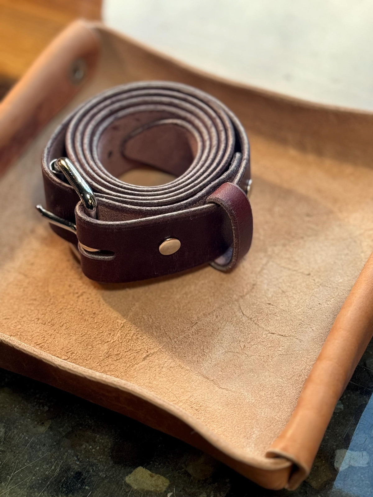 A dark brown leather belt with a silver-tone buckle, resting on a tan leather surface.